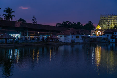 Trivandrum, India - February 28, 2023: The illuminated gopuram of Sree Padmanabhaswamy Temple is visible behind traditional buildings and the sacred temple tank at dusk, with reflections on the water.のeditorial素材