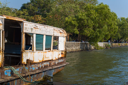 Kochi, India - February 27, 2023: A rusty and dilapidated ferry with broken windows is docked at the edge of the water, with a stone walkway, trees, and people visible along the shoreline.のeditorial素材