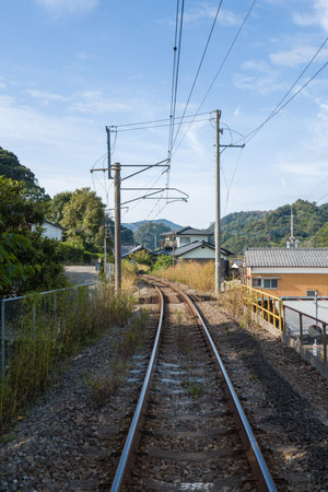 Arita, Japan - November 9, 2024: The image shows a single railway track with overhead electric lines curving through a residential neighborhood, with houses on the right, dense greenery on the left, and mountains in the background under a partly cloudy skのeditorial素材
