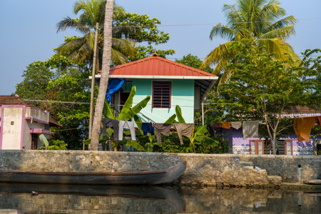 Alappuzha, India - March 7, 2023: Laundry is hung on clotheslines in front of colorful residential houses with palm trees, banana plants, and a stone embankment beside a canal, with a wooden boat moored along the water.のeditorial素材