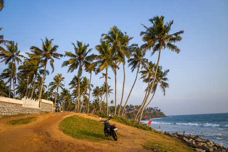 Varkala, India - March 2, 2023: A motorbike is parked on a dirt path lined with palm trees near a stone wall, overlooking Odayam Beach with waves and people visible in the water under a clear sky.のeditorial素材
