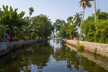 Alappuzha, India - March 7, 2023: A canal with stone embankments is lined with palm trees and dense vegetation, with small boats moored along the water and laundry hanging on the left side under a clear sky.のeditorial素材