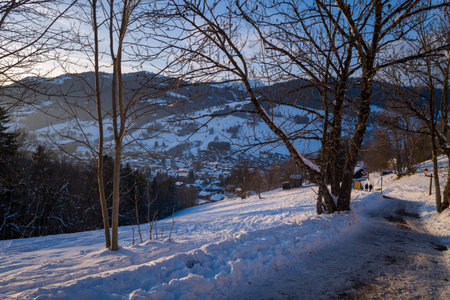 Megeve, France - January 30, 2023: A snow-covered path lined with bare trees descends towards the village of Megeve, with houses and mountains visible in the background at sunset.のeditorial素材