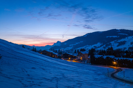Megeve, France - January 30, 2023: A snow-covered hillside descends towards a village street with illuminated lamps and scattered houses, with mountain silhouettes visible in the background at dusk.のeditorial素材