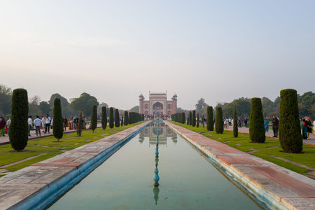 Agra, India - March 11, 2024: The main red sandstone entrance gate of the Taj Mahal complex is seen in the distance, reflected in the central pool, with rows of cypress trees and groups of people walking along the Mughal garden pathways under a clear mornのeditorial素材