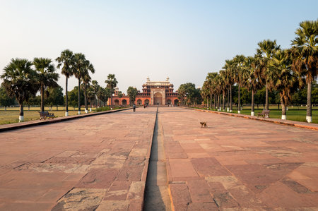 Agra, India - March 12, 2024: The image shows a broad stone pathway lined with palm trees leading to the main entrance gate of Akbars Tomb, with benches, green lawns, a few people, and a monkey visible under a clear sky.のeditorial素材