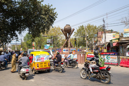 Mathura, India - March 13, 2024: Motorcycles, an auto rickshaw, pedestrians, and a large metallic sculpture are visible at a busy intersection in Mathura, with commercial signage, traffic lights, and trees in the background during daylight.のeditorial素材