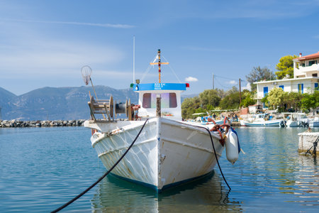 Port de Vathi, Greece - September 3, 2024: Close-up view of a fishing boat moored at the quay with residential houses, other small boats, and mountains visible in the background under a clear blue sky.のeditorial素材
