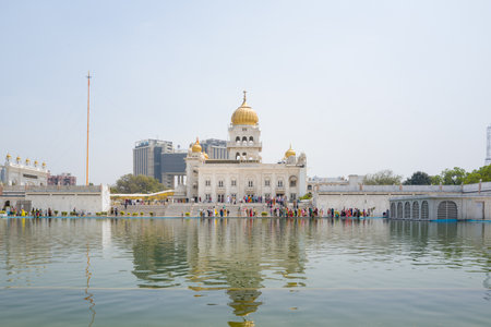 Delhi, India - March 29, 2024: The white marble building of Gurudwara Sri Bangla Sahib with golden domes is seen across the sarovar, with groups of people gathered along the waters edge under a clear sky.のeditorial素材