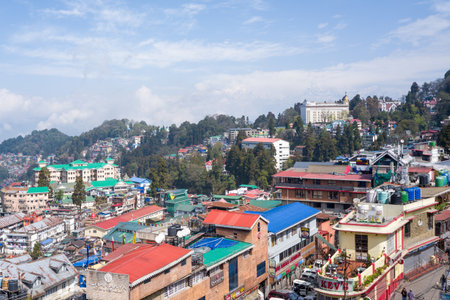 Darjeeling, India - March 26, 2024: An elevated view shows the cityscape of Darjeeling with multi-story buildings, colorful rooftops, shops, water tanks, signage, and distant hills under a partly cloudy sky.のeditorial素材