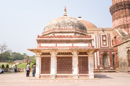 Delhi, India - March 30, 2024: The image shows the front view of Imam Zamins Tomb with its white marble dome, red sandstone lattice windows, and several people standing nearby at the Qutub Minar complex.のeditorial素材