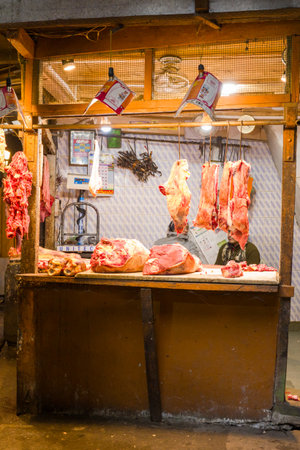 Darjeeling, India - March 26, 2024: Cuts of raw meat hang from hooks and are laid out on a wooden counter inside a small butcher shop, with two people partially visible behind the counter under bright lights.のeditorial素材