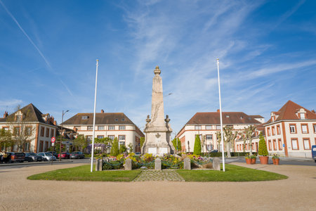 Unknown location - April 13, 2024: The image shows a stone war memorial monument at the center of Place Aristide Briand, flanked by two empty flagpoles, with flower beds and historic buildings in the background under a clear blue sky.のeditorial素材