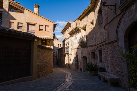Alquezar, Spain - May 6, 2024: View of a narrow cobblestone street lined with historic stone and brick buildings, iron-barred windows, balconies, and a stone bench in the old town center of Alquezar under clear daylight.のeditorial素材