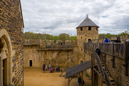 Chateau de Guedelon, France - April 20, 2024: The image shows people in the courtyard and on a wooden staircase and walkway inside Chateau de Guedelon, with stone walls, a round tower, and cloudy sky visible.のeditorial素材