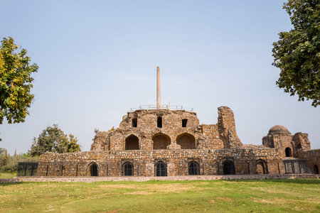 Delhi, India - March 29, 2024: The image shows the Topra Ashokan pillar standing atop a multi-level stone structure with arched openings and iron gates at Feroz Shah Kotla Fort, with a grassy lawn and trees visible in the foreground under a clear sky.のeditorial素材