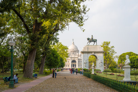 Calcutta, India - March 23, 2024: The image shows a gravel path lined with mature trees and lampposts leading towards an equestrian statue on an arch and the Victoria Memorial, with people walking and sitting on benches in the surrounding gardens.のeditorial素材