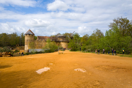 Chateau de Guedelon, France - April 20, 2024: Several people are walking and standing on a large open dirt area in front of the stone towers and partially scaffolded walls of Chateau de Guedelon, with surrounding trees and a cloudy sky.のeditorial素材