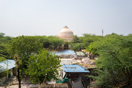 Delhi, India - March 30, 2024: The image shows a large domed tomb in the Mehrauli Archaeological Complex, partially hidden by dense green trees and surrounded by temporary structures with metal roofs in the foreground.のeditorial素材