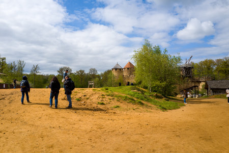 Chateau de Guedelon, France - April 20, 2024: Several people are seen walking and standing on a dirt path with the stone towers, wooden crane, and surrounding greenery of Chateau de Guedelon visible in the background under a partly cloudy sky.のeditorial素材
