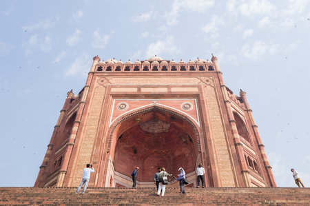 Fatehpur Sikri, India - March 12, 2024: The image shows a low angle view of the Buland Darwaza, the monumental main entrance gate to the Jama Masjid mosque at Fatehpur Sikri, with several people visible on the wide stone steps under a partly cloudy sky.のeditorial素材