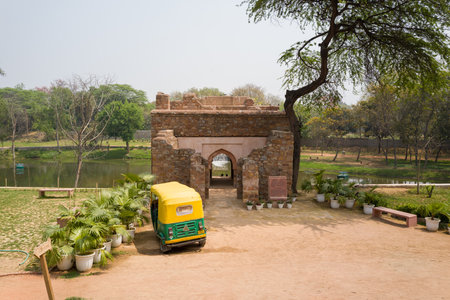 Delhi, India - March 30, 2024: The image shows the Boat House structure at the Mehrauli Archaeological Complex with a yellow-green auto rickshaw parked in front, potted plants, benches, and a pond surrounded by trees in the background.のeditorial素材