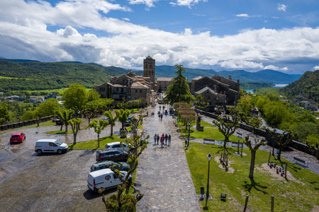 Unknown location - May 6, 2024: Elevated view of a cobblestone street with people walking, parked cars, trimmed trees, stone buildings, and a prominent church tower, with green hills and a lake in the background.のeditorial素材