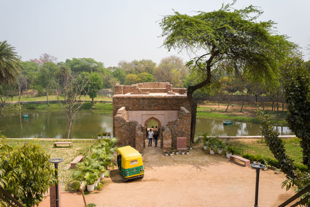 Delhi, India - March 30, 2024: The image shows the Boat House structure at the Mehrauli Archaeological Complex with two people standing at the entrance, a yellow-green auto rickshaw parked in front, potted plants, and a pond in the background surrounded bのeditorial素材