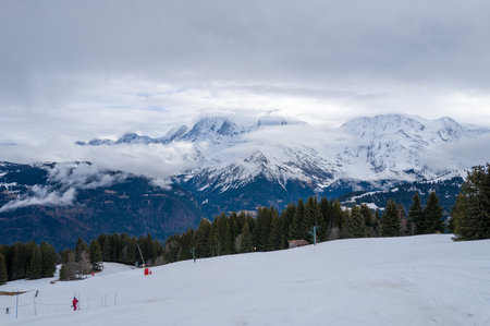 Megeve, France - February 8, 2024: A snow-covered ski slope with several skiers is visible in the foreground, bordered by pine trees, with the Mont Blanc massif partially covered by clouds in the background under an overcast sky.のeditorial素材