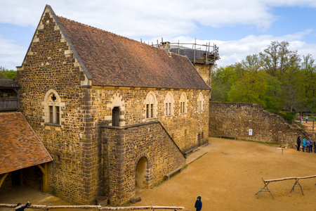 Chateau de Guedelon, France - April 20, 2024: The image shows the main stone building of Chateau de Guedelon with arched windows, a tiled roof, and an adjacent courtyard with several people and construction materials visible under a partly cloudy sky.のeditorial素材