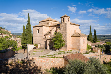 Alquezar, Spain - May 6, 2024: Exterior view of the historic Church of San Miguel with stone walls, cypress trees, and adjacent village buildings under a partly cloudy sky.のeditorial素材