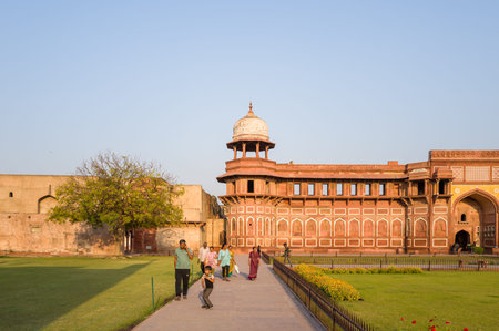 Agra, India - March 11, 2024: Several people are seen walking and standing on a paved pathway bordered by manicured lawns and flower beds in front of the red sandstone facade and corner pavilion of Jahangir Palace at Agra Fort under a clear sky.のeditorial素材
