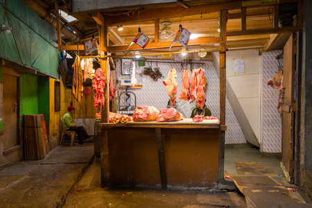 Darjeeling, India - March 26, 2024: Cuts of raw meat hang from hooks and are displayed on a counter inside a small butcher shop, with two people visible, one sitting to the side and another partially behind the counter.のeditorial素材