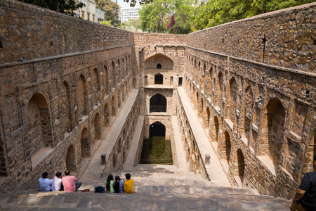 Delhi, India - March 29, 2024: Several people are seated on the stone steps overlooking the arched corridors and central well of Agrasen ki Baoli, a historic stepwell with multi-level stone architecture and water at the bottom, under daylight.のeditorial素材