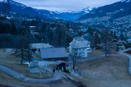 Megeve, France - February 7, 2024: An elevated view shows buildings, winding roads, and bare ground in Megeve village, with the surrounding mountains visible in the background and little to no snow on the landscape.のeditorial素材