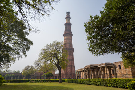 Delhi, India - March 30, 2024: The image shows the Qutub Minar rising behind trees and a green lawn, with adjacent stone ruins of the Qutub Minar archaeological complex visible on the right under a clear sky.のeditorial素材