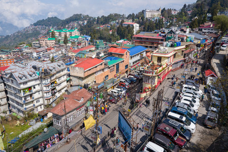 Darjeeling, India - March 26, 2024: An elevated view shows the cityscape of Darjeeling with multi-story buildings, colorful rooftops, shops, parked vehicles, pedestrians, and distant hills under a partly cloudy sky.のeditorial素材