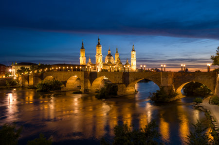 Zaragoza, Spain - May 11, 2024: The image shows the Basilica of Nuestra Senora del Pilar with its towers and domes illuminated at twilight, behind the lit Stone Bridge spanning the Ebro River with reflections on the water.のeditorial素材