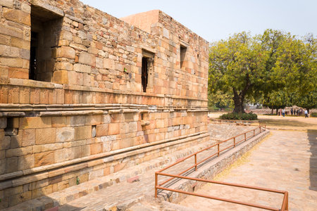 Delhi, India - March 30, 2024: The image shows a section of a historic stone wall with rectangular windows and a ramp with metal railings at the Qutub Minar complex, with trees and several people visible in the background.のeditorial素材