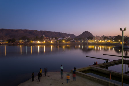 Pushkar, India - March 18, 2022: People are seen standing and sitting on the ghats by Pushkar Lake at dusk, with reflections of illuminated town buildings and hills visible across the calm water.のeditorial素材