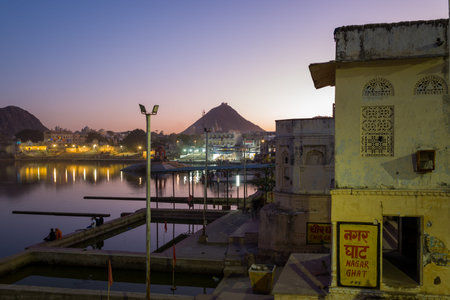 Pushkar, India - March 18, 2022: Nagar Ghat is seen at dusk along Pushkar Lake, with people sitting by the water, illuminated town buildings, and hills visible in the background.のeditorial素材