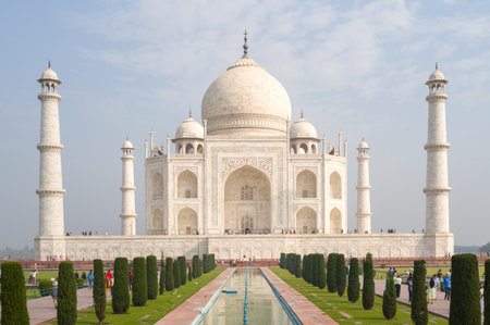 Agra, India - March 11, 2024: The Taj Mahal is seen from the front with its marble dome, four minarets, and main entrance, as groups of people walk and stand along the central Char Bagh garden and reflecting pool under a partly cloudy sky.のeditorial素材