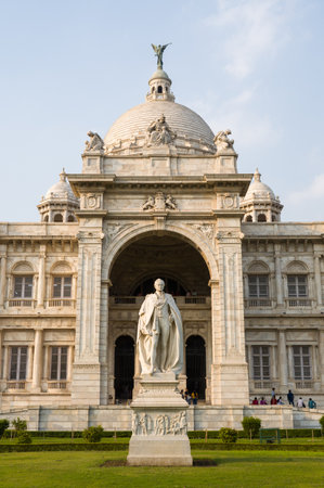 Calcutta, India - March 23, 2024: The front entrance of Victoria Memorial is shown with its marble dome, neoclassical architecture, and a statue of Lord Curzon on the lawn, while visitors are visible near the entrance under a clear sky.のeditorial素材