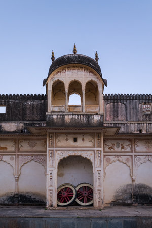 Pushkar, India - March 18, 2022: The exterior wall of Sri Rangnath Swamy Temple features a domed balcony, ornate painted arches, faded animal motifs, and two large wooden cart wheels placed under a central archway at dusk.のeditorial素材