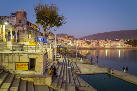 Pushkar, India - March 18, 2022: People are seen sitting and walking on the steps of Jagannath Ghat by Pushkar Lake at dusk, with illuminated buildings, signage in Hindi, and hills visible in the background.のeditorial素材