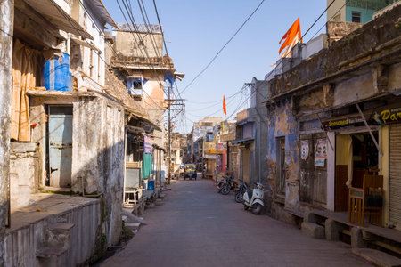 Bundi, India - March 19, 2022: A narrow street lined with weathered residential and commercial buildings, visible signage, parked motorcycles and scooters, overhead power lines, and orange flags during daylight.のeditorial素材