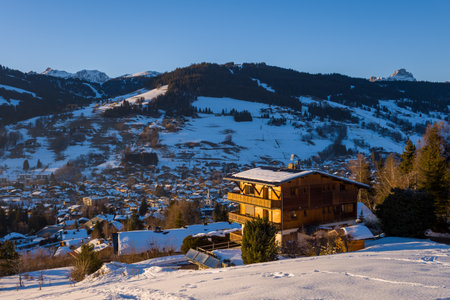 Megeve, France - January 23, 2022: A wooden chalet with a snow-covered roof is seen on a hillside overlooking the town of Megeve, with snow-covered slopes, forested mountains, and clear blue sky in the background.のeditorial素材