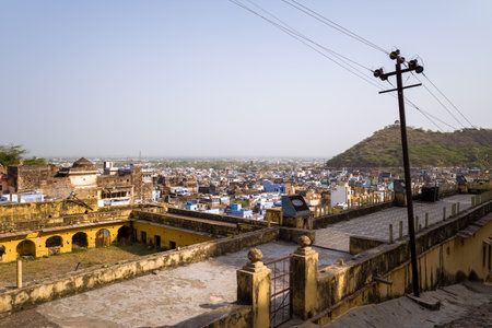 Bundi, India - March 20, 2022: The image shows an elevated view over Bundi with historic yellow buildings in the foreground, a dense cluster of city structures, a hill with greenery, and electrical poles and wires under a clear sky.のeditorial素材