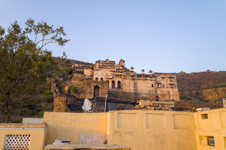 Bundi, India - March 19, 2022: The Garh Palace complex is seen on a hillside with weathered yellow walls, arched gateways, and multiple domed pavilions, with foreground rooftops and a tree under a clear sky.のeditorial素材
