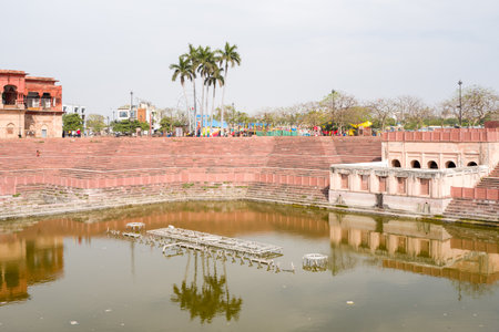 Lucknow, India - March 16, 2024: The image shows a large stepwell with red sandstone terraces, a pool of water, and a historic building near the Hussainabad Clock Tower, with palm trees and people visible in the background under daylight.のeditorial素材